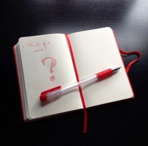 An open notebook with the words "Think of a name" and a big question mark written in red, with a red pen lying horizontally across the page.