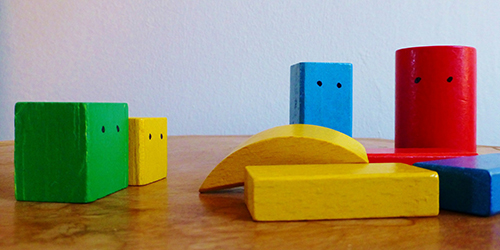 A colourful (green, yellow, blue, red) set of children's wooden building blocks of diffferent shapes and sizes, with dots for eyes painted on them. It conveys building a team.