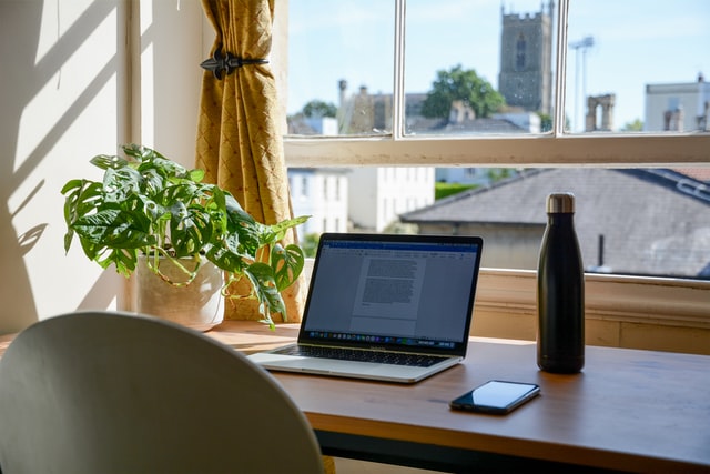 Office desk by a window with a pot plant, laptop, water bottle and mobile phone on it.