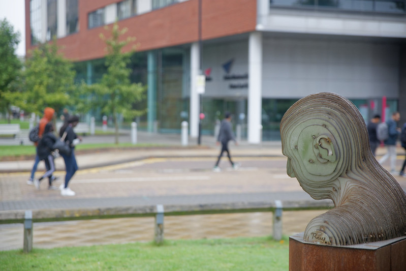 Students walking down a road with a university builiding in the background. A statue of a profile of a human head is in the foreground on the right-hand side.